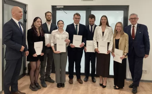 Grupa siedmiu młodych osób trzymających certyfikaty. Są ubrani w formalne stroje i stoją przed szklanymi drzwiami. Towarzyszą im dwaj mężczyźni w garniturach.