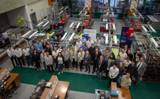 Liczna grupa osób pozująca do zdjęcia w laboratorium, w którym znajduje się wiele różnych stanowisk z maszynami innymi urządzeniami
