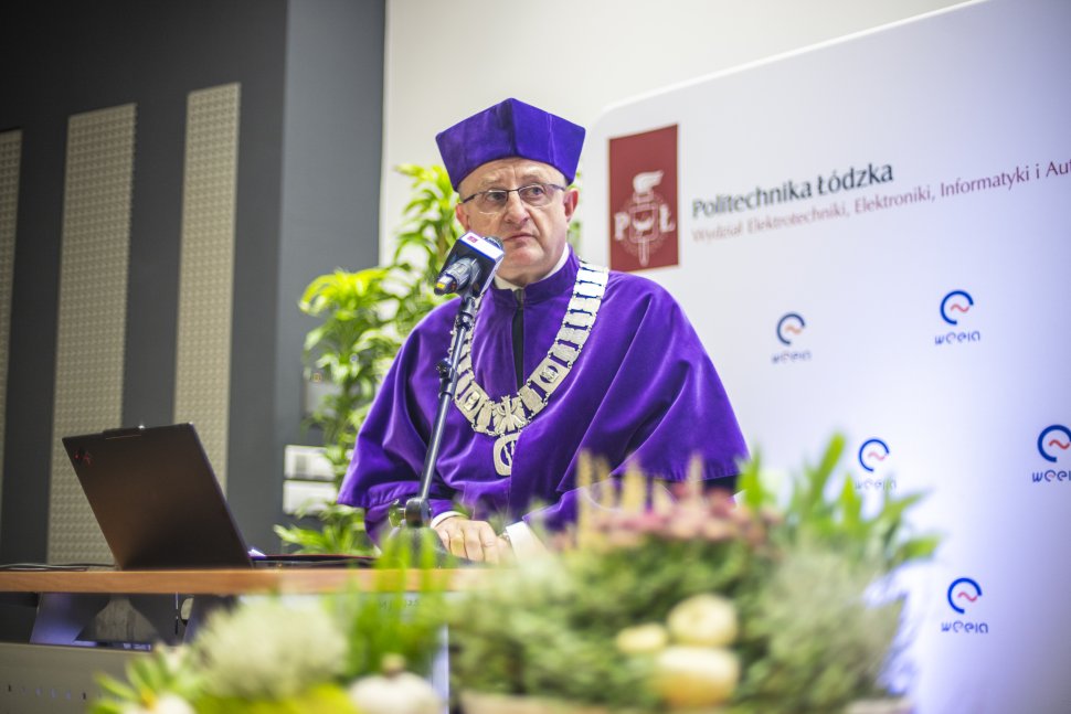 Dziekan wydziału w todze, z łańcuchem na klatce piersiowej, mówi do mikrofonu. W tle ścianka z logo WEEIA. Przed dziekanem laptop. Na pierwszym planie rozmyta zielona dekoracja roślinna.